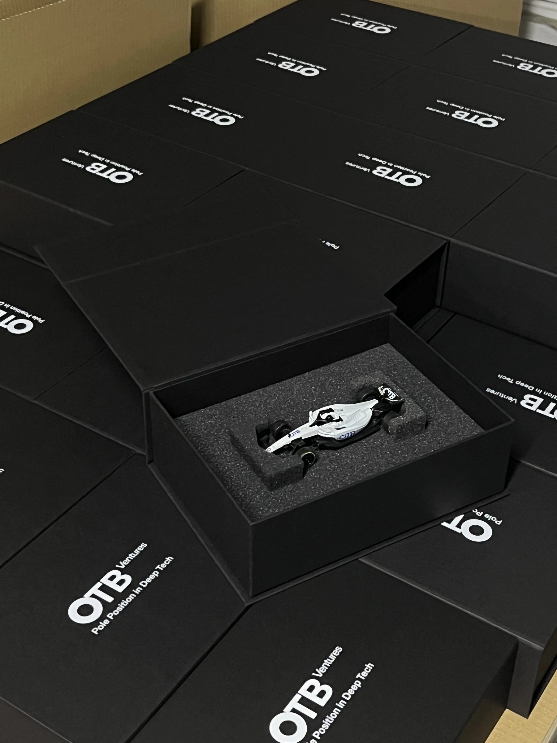 Model bolidu OTB Ventures w otwartym opakowaniu prezentowym, na tle serii zamkniętych pudełek